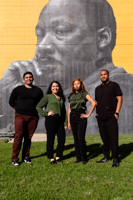 Four young Brown people stand tall in a line, in sun so their shadows fall behind them, the women in the middle with one arm on a hip, in front of a detailed grayscale mural of Martin Luther King Jr on a yellow background.