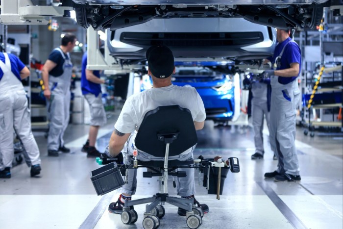 A man sits on an office chair with his back to the camera watching people on the assembly line