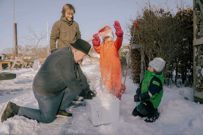 An older man in hat packs snow into a bucket while a younger man and two children watch on