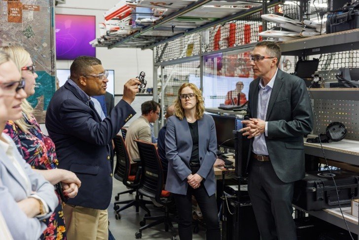 Two men in safety goggles conversing while one holds a piece of technology up.