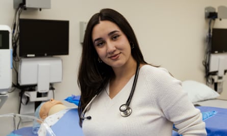 A young light-skinned woman with long dark hair parted in the middle, a long-sleeved white shirt, and a stethoscope around her neck, posing and looking at the camera smiling with her head tilted to the right.