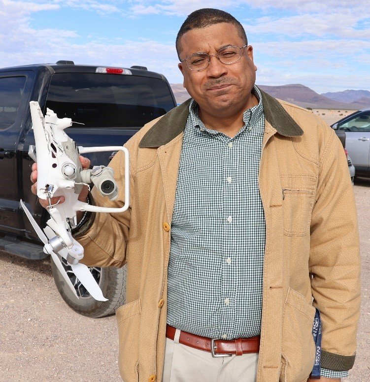 Large full-body shot of Deputy Administrator Rose. In his rand hand he holds up a white destroyed asset over a desert landscape.