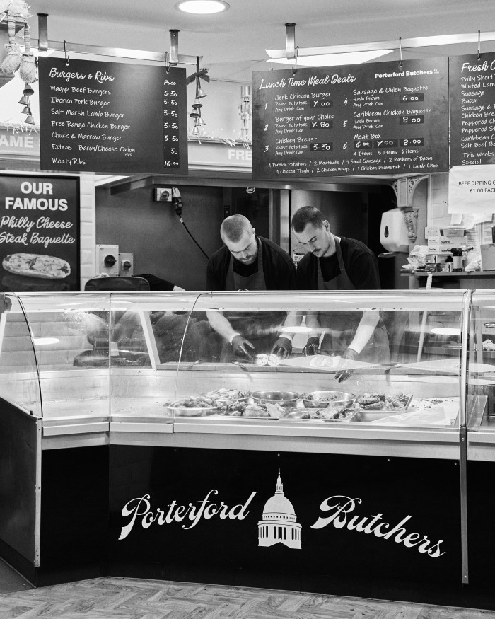 A black and white photo of Porterford butcher, with the menu written in chalk on blackboards behind two male employees behind the counter