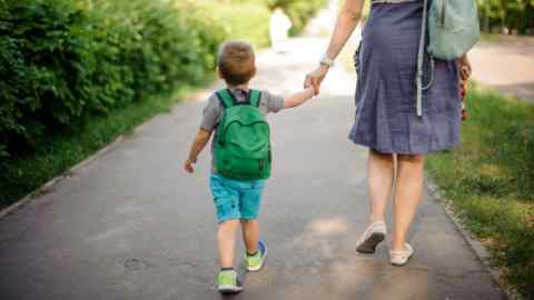 Back view of mother walking down the street with a little son with a backpack on sunny summer day