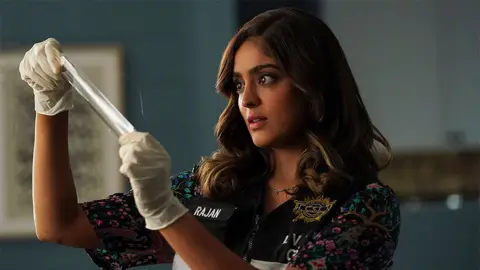 Getty Images An image of Mandeep from the TV show CSI: Vegas, in which she is wearing gloves, a police vest and is looking at a piece of white tape.