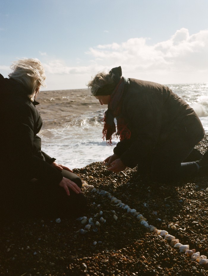Two women kneeling on a pebbly beach arrange shells in a line with the sea in the background