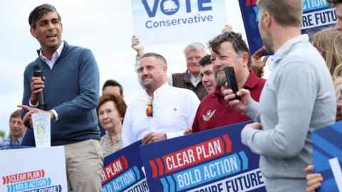 Rishi Sunak at a campaign event, with people holding Conservative party placards in the background