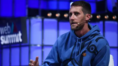 Getty Images A man speaking on-stage at an event. He's wearing a blue hoody and has a smart beard and hairstyle