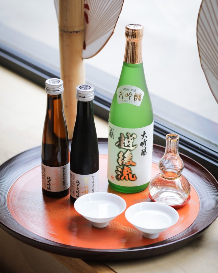 A large green bottle, two smaller brown bottles and a carafe of sake – alongside small sake bowls – on a circular red tray at Roketsu