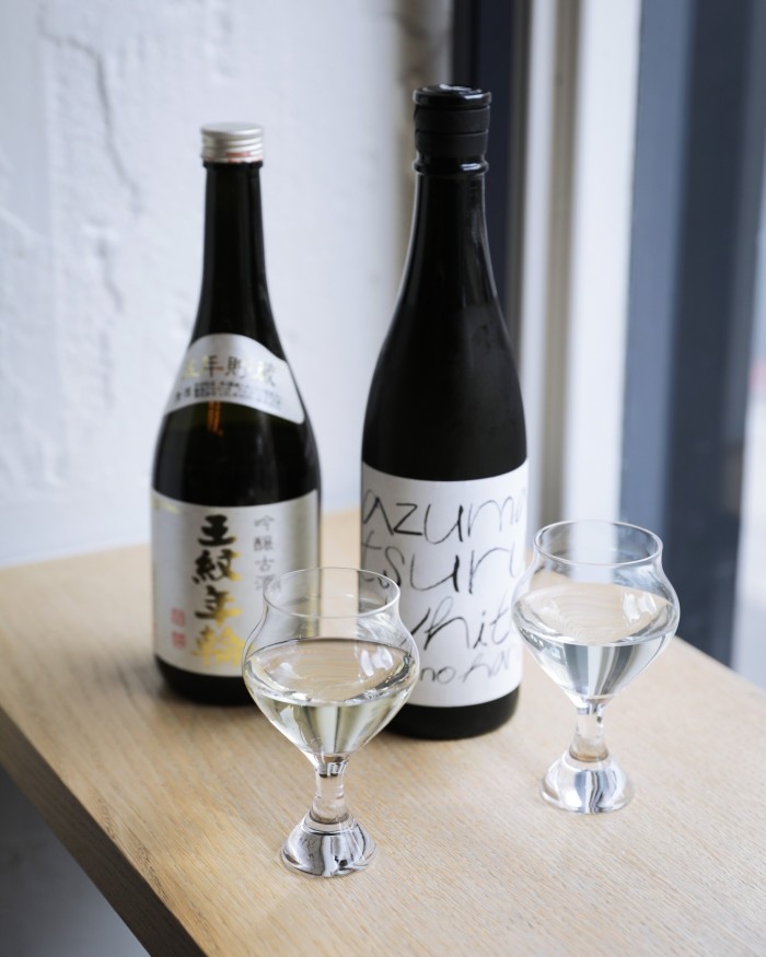 Two bottles and two glasses of sake on a pale-wood counter at Sake Collective