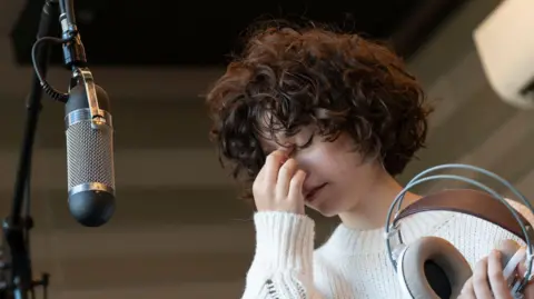 Getty Images A stock image of a female vocal artist hand covering her face, her eyes closed, standing in front of a large microphone on a stand