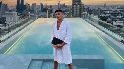 Instagram Samy Daim wearing a white dressing gown and holding what appears to be an iPad. He is standing by an infinity pool with a city skyline and sunset behind. 