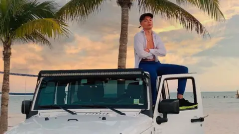 instagram Samy Daim sitting on top of a white 4x4 vehicle parked beside a beach with  palm trees in the background. Mr Daim is wearing an unbuttoned white shirt with blue trousers and cap.