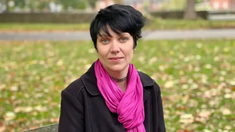 BBC/BRANWEN JEFFREYS Rebekah looks at the camera sitting on a wooden bench in a park. She has short black hair and is wearing a dark pink scarf, a black choker and a black jacket