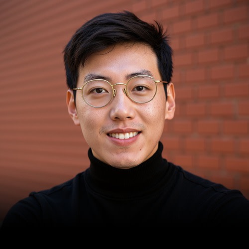 A portrait of a man with neatly styled hair smiling for the camera. He’s wearing round glasses and a turtleneck sweaters. In the background is a red brick wall