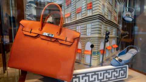 An orange Hermès handbag is prominently displayed in a boutique window against a backdrop featuring a stylised model of a Parisian building. A pair of blue sandals is also on display