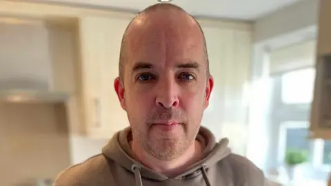 James Gilmartin Close up of James Gilmartin who has a shaved head, wearing a light brown hoody, standing in his kitchen