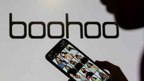 A woman poses with a smartphone showing the Boohoo app in front of the Boohoo logo