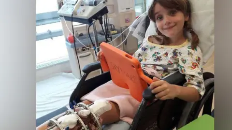 Dean Stalham A young girl lying in a hospital bed wearing pyjamas and looking at an ipad with a metal frame on her left leg.