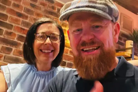 Richard Donaldson Man with ginger beard, wearing a bunnet, wearing a dark top and holding his thumb up, smiling, with a smiling woman with side-parted dark hair and glasses, wearing a short-sleeved light blue blouse, with her hand round his upper arm.