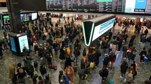 PA Media Passengers standing on a railway concourse in front of large departure screens