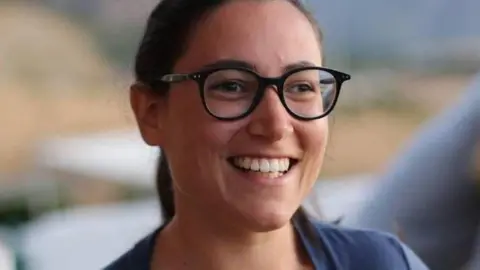 Martino Brizio Sofia Brizio, a young woman with brown hair, brown eyes, wearing dark framed glasses and wearing a mid blue top smiles looking slightly off camera, the background is blurred.