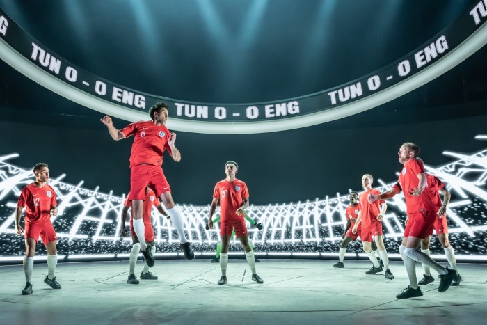 A group of men in England football strips stand on a stage which is designed to look like a football stadium. One of the men is jumping into the air to header a ball, while the others look on