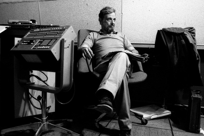 A black and white image of a man with greying hair and a beard sitting back on a chair in a recording studio of some kind. He has a newspaper on his knee and is looking to his left, with a slight smile