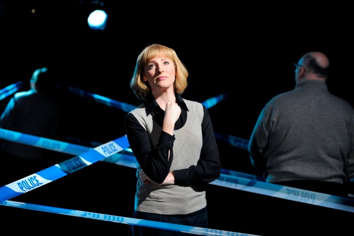 A woman with shoulder-length blond hair is standing on a stage between several strips of police incident tape. She has a sad, pensive expression. Two men can be seen behind her