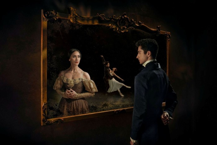 A man and a woman stand and look at one another from either side of an ornate frame. Both are in period costume. To one side of the woman, a couple are dancing