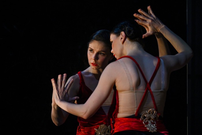 A woman wearing a red sleeveless dress and matching lipstick leans against a mirror, in which she is reflected. She has a worried expression