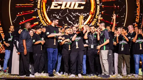 Luc Bouchon A large group of young men wearing black t-shirts with "Champions" printed on them stand on a stage littered with pieces of confetti as one member at the front of the group triumphantly holds up a tall golden trophy amid the scenes of celebration. Behind them an "EWC: Esports World Cup" logo is visible. 