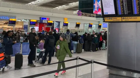 BBC People in coats and with suitcases stand in a line in a Heathrow Airport terminal. A woman in a green coat and green trousers walks in the opposite direction