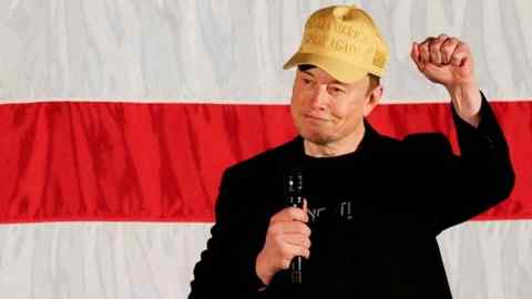 Elon Musk, wearing a cap that reads "Make America Great Again," gestures with his fist raised while holding a microphone