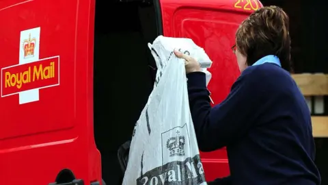 PA Media With her back to the camera a shorthaired woman wearing a navy sweater lifts a grey sack into the back of red Royal Mail van. 