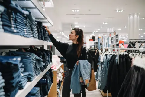 Getty Images Young woman with brown hair in clothing store, looking at shelves stacked with jeans.