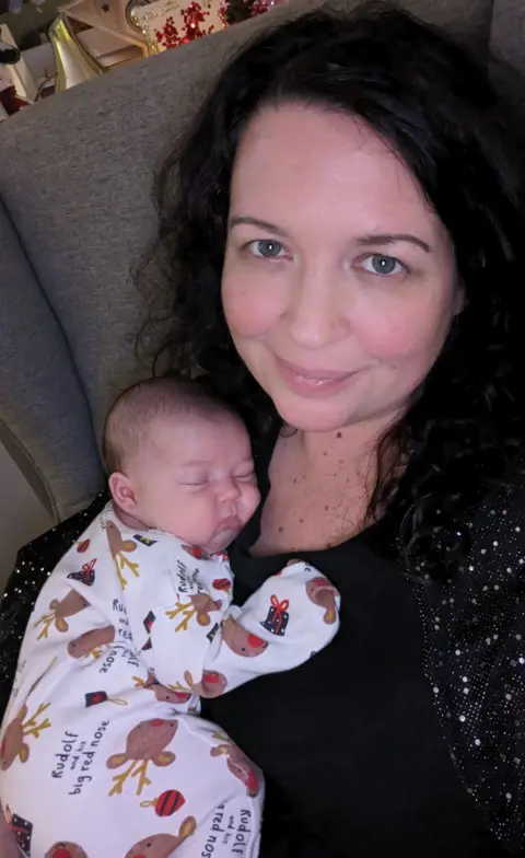 Adele Darlington Adele sits on a grey sofa, smiling and facing the camera, cuddling her baby daughter Jasmine who is wearing a Christmas-themed babygro