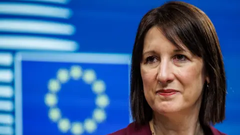 EPA UK Chancellor Rachel Reeves stands in front of a blue and white EU flag She wears a burgundy suit and her fringe is swept to the side. 