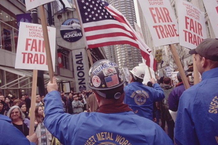 A group of people protest in the street waving the US flag and banners with Fair Trade written on them