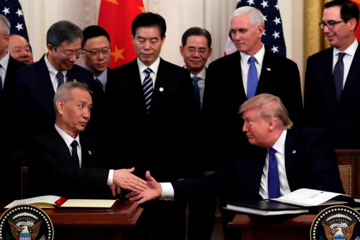 A group of men in suits stand looking at two men seated at a table shaking hands