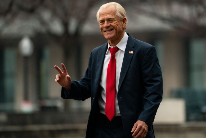 A smiling grey-haired man in a dark suit and red tie, in an outside setting, waves his right hand